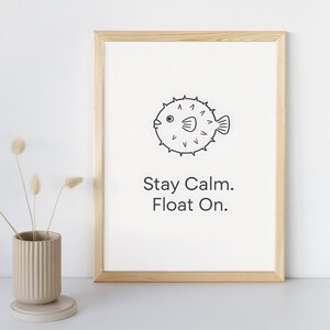 May include: A framed print with a light wood frame. The print features a black and white illustration of a pufferfish and the text "Stay Calm. Float On." A beige ceramic vase with dried flowers is in the foreground.
