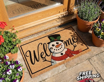 May include: A rectangular doormat with a snowman design and the word "Welcome" in black script. The snowman wears a black top hat with holly, a red scarf, and has a carrot nose. The mat is made of a natural fiber material.