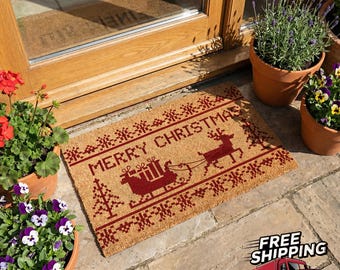 Könnte beinhalten: Eine rechteckige Fußmatte mit festlichem "MERRY CHRISTMAS"-Design. Die Matte zeigt ein Rentier, das einen Schlitten mit Geschenken zieht, alles in tiefroter Farbe auf beigem Hintergrund. Die Matte liegt vor einer Holztür.