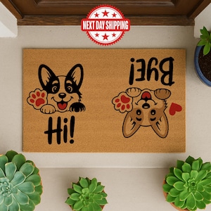 Puede incluir: Una alfombra rectangular con un diseño de perro corgi. Un corgi está de pie con la palabra "Hi!" debajo, y el otro está boca abajo con la palabra "Bye!" encima. La alfombra es marrón con detalles en negro y rojo.