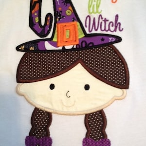 May include: Embroidered design of a girl wearing a purple and orange witch hat with a spiderweb and stars. The text "Mommy's lil Witch" is above the girl's head. The design is on a white background.