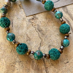 Op de afbeelding: Een armband met turquoise lavasteen kralen, houten kralen en zilverkleurige metalen kralen. De kralen zijn geregen op een elastisch koord. De achtergrond is een houten oppervlak.