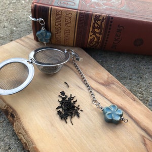 May include: A silver tea infuser with a blue ceramic flower charm on a chain. The infuser is open and a small pile of loose leaf tea is on the wooden surface.