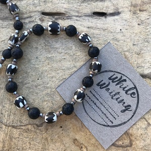 May include: Black and white patterned beads with black lava rock beads on a string. The beads are strung on a silver cord. The necklace is on a wooden surface. The text "while waiting" is on a brown card.