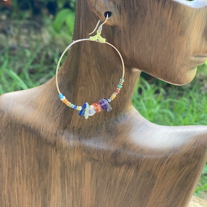 May include: Silver hoop earrings with a rainbow of small gemstone beads and a larger gemstone charm. The charm features a variety of colors, including red, orange, yellow, green, blue, indigo, and violet.
