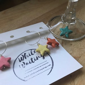 May include: Four colorful star-shaped glass charms on silver hoops. The charms are turquoise, yellow, orange, and pink. The charms are labeled "While Waiting..." and "www.whilewaitingshop.com".