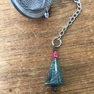 May include: A stainless steel tea infuser with a chain and a decorative charm. The charm is a green, cone-shaped stone with a red star on top. The infuser is designed for steeping loose leaf tea.