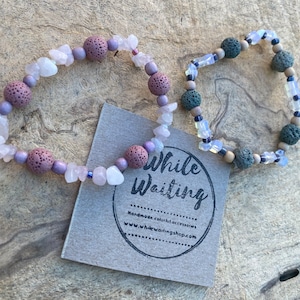 May include: Two bracelets made with lava rock beads, rose quartz, and opal beads. The bracelets are on a wooden surface with a business card that says "While Waiting" and "Handmade colorful accessories www.whilewaitingshop.com".