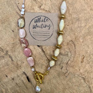 May include: A handmade necklace featuring a mix of pink, cream, and gold beads. The necklace includes large, irregular pink stones, cream-colored paper beads, and gold-colored metal accents. A gold toggle clasp and a pearl pendant complete the design. The tag reads "While Waiting".