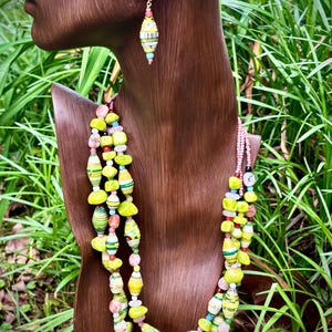Puede incluir: Un collar de varias hebras y un pendiente a juego con una variedad de cuentas en tonos verde lima, amarillo, rosa y blanco. Las cuentas incluyen formas cilíndricas tipo papel, piedras redondas lisas y pequeños detalles translúcidos.
