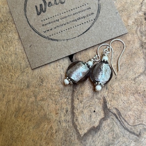 May include: Silver-toned dangle earrings with oval, textured, dark gray beads and small white accents. The earrings are displayed on a wooden surface next to a brown card with the text "While Waiting".