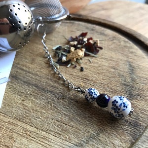 以下が含まれることがあります： 銀製の茶こしにチェーンと青と白のセラミックビーズのチャームが付いています。茶こしは木製の表面にあり、その周りに茶葉が散らばっています。