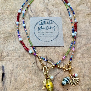 May include: Two beaded necklaces with gold clasps and charms. One necklace features a yellow and green paper bead charm with a gold fish charm. The other has a black and white bead charm and a gold bee charm. The necklaces are on a wooden surface.