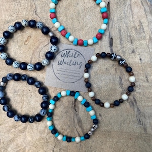 May include: Five beaded bracelets in various designs and colors are displayed on a wooden surface. The bracelets feature beads in turquoise, red, black, white, and silver. A small card with the text "While Waiting" is also visible.