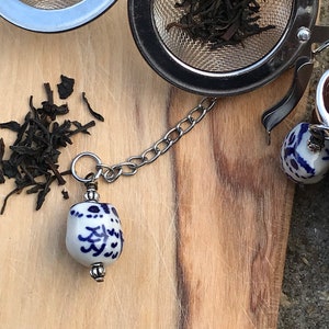 May include: A silver tea infuser with a chain and a blue and white ceramic bead. The infuser is filled with loose leaf tea.