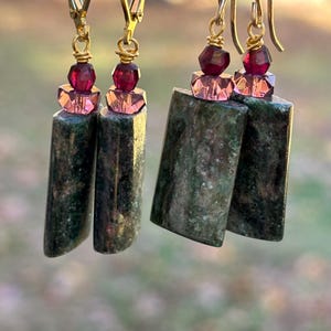 May include: Gold-tone dangle earrings with green stone rectangles, accented by pink and burgundy beads. The earrings hang from gold-colored ear wires, showcasing a natural, earthy aesthetic. The earrings are a unique accessory.