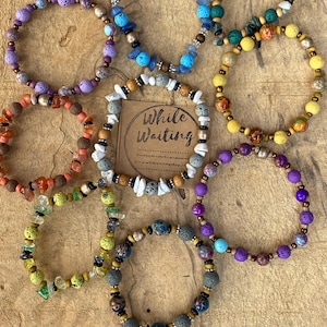 May include: Assortment of colorful bracelets made with lava rock beads, wood beads, and gemstone beads. The bracelets are arranged on a wooden surface with a sign that reads "While Waiting".