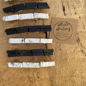 May include: Set of six hair clips made of white howlite and black lava rock. The clips are arranged in alternating colors and are displayed on a wooden surface. The image also includes a small business card with the text "While Waiting" and a website address.