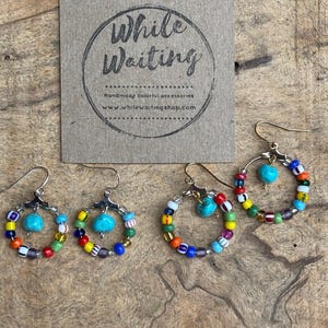 May include: Three pairs of handmade hoop earrings with colorful glass beads and turquoise accents. The earrings are displayed on a wooden surface next to a brown card with the text "While Waiting".