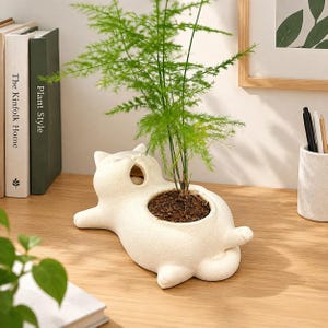 Puede incluir: Una maceta de cerámica color crema con forma de gato recostado y superficie texturizada. El gato está tumbado de lado con la cabeza apoyada cerca de una abertura circular que contiene una pequeña planta verde de hojas finas y plumosas.