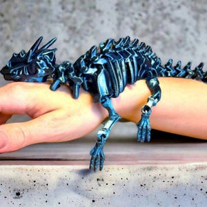 Puede incluir: Una pulsera de dragón articulada de color azul oscuro. El diseño esquelético del dragón presenta una cabeza, una columna vertebral y extremidades detalladas. La pulsera se muestra en una muñeca, mostrando su construcción flexible e intrincada. El color del dragón tiene un brillo metálico.