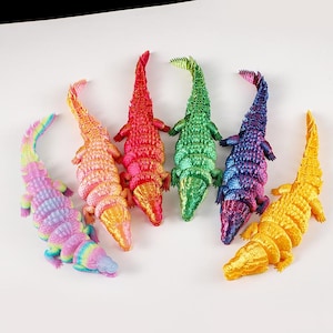 May include: Six colorful alligator figurines in a variety of hues, including pink, orange, red, green, blue, and yellow. Each alligator has a textured, scaly design and is lying flat. The figurines are likely decorative items or toys.