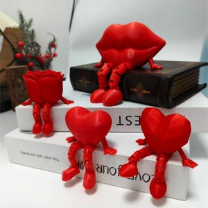Puede incluir: Una colección de figuras rojas impresas en 3D, que incluyen una rosa, labios y corazones, cada una con piernas y pies articulados. Las figuras están dispuestas sobre libros, creando una exhibición lúdica y caprichosa.