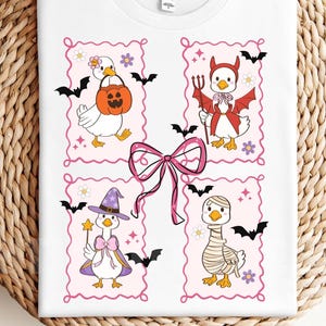 May include: White t-shirt featuring four cartoon geese in Halloween costumes. One goose carries a pumpkin, another is a devil, a third is a witch, and the last is a mummy. Pink frames, bats, and flowers surround each goose.