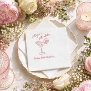May include: A square white paper napkin featuring a red line drawing of a martini glass with a lemon slice and the text "a tajini bit older" above the glass and "Emily's 30th Birthday" printed in red ink below the illustration.