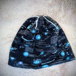 Könnte beinhalten: Eine dunkelgraue Mütze mit futuristischem Design. Sie zeigt Binärcode, leuchtende blaue Kreise und die Wörter "EXERCISE" und "LOADING...". Die Mütze hat einen weißen Kordelzugverschluss.