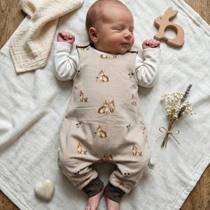 Könnte beinhalten: Ein schlafendes Baby in einem beigen Overall mit Hasenmuster, weißen Langarmärmeln und braunen Bündchen. Das Baby liegt auf einer weißen Decke mit einem Holz-Hasen-Spielzeug, einem herzförmigen Stein und Trockenblumen.