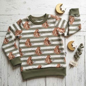 Könnte beinhalten: Ein langärmliges Sweatshirt mit olivgrünen und weißen Streifen und einem sich wiederholenden Muster aus braunen Bären. Die Bündchen, der Halsausschnitt und der Saum sind einfarbig olivgrün. Der Text "The Magh... Back" ist aufgedruckt.