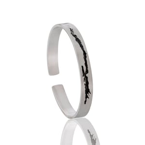 May include: A silver bracelet with a cut-out design of a lake or island. The bracelet is open and has a smooth, polished finish.