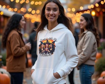 May include: A white hooded sweatshirt featuring a graphic of three witches with the text "Partying with My Witches". The sweatshirt has a front pocket and a drawstring hood. The image is set against a blurred outdoor background.