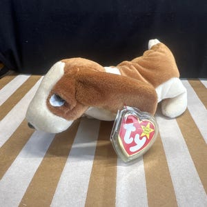 Puede incluir: Un peluche de perro Basset Hound con pelo marrón y blanco, tumbado sobre una superficie a rayas. Una etiqueta en forma de corazón con el logotipo "Ty" está adjunta al juguete.