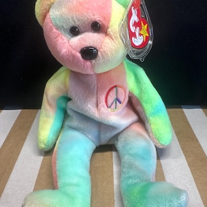 May include: A colorful plush bear with a peace sign on its chest. The bear features a tie-dye pattern in shades of pink, yellow, green, and blue. It has a black nose and eyes, and a tag attached to its ear.