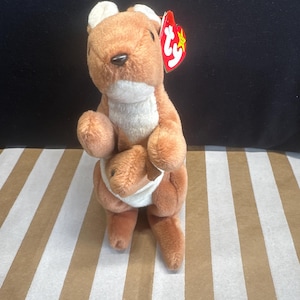 May include: A plush kangaroo toy in shades of brown and white, with a small joey in its pouch. The toy has a red tag with a yellow star. The kangaroo is standing on a brown and white striped surface.