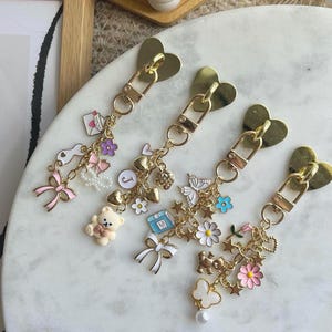 May include: Four gold keyrings with heart-shaped clasps. Each keyring has a selection of charms, including bows, flowers, stars, a teddy bear, and a butterfly. The charms are in pastel colours with gold accents.