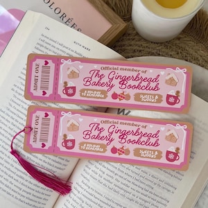 Puede incluir: Dos marcapáginas rosas y beige con el texto "Official member of The Gingerbread Bakery Bookclub" e ilustraciones navideñas. Una borla rosa cuelga de un marcapáginas. Los marcapáginas están sobre un libro abierto.