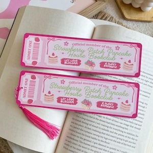 Puede incluir: Dos marcapáginas rosas de Strawberry Patch Pancake House Bookclub con borlas rosas sobre un libro abierto. Los marcapáginas presentan texto e ilustraciones de fresas y panqueques.