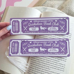 Op de afbeelding: Twee paarse bladwijzers met witte tekst en een ticketontwerp. De bladwijzers lezen "Official member of the Quicksilver Book Club" en bevatten een vosillustratie. Elke bladwijzer heeft een witte kwast bevestigd. De bladwijzers liggen op een open boek.
