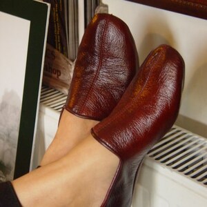 May include: A pair of burgundy leather slippers with a textured finish. The slippers are worn on a person's feet, resting on a white radiator.