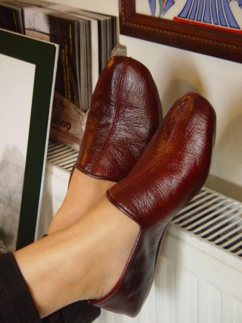 May include: A pair of burgundy leather slippers with a textured finish. The slippers are worn on a person's feet, resting on a white radiator.