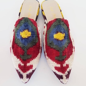 Puede incluir: Un par de mules de terciopelo coloridas con punta. Los zapatos presentan un diseño de flores rojas, blancas, azules y amarillas. El interior del zapato es amarillo y tiene el texto "Juju Shoes".
