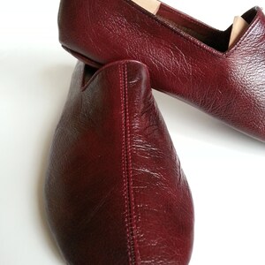 May include: A pair of burgundy leather slippers with a pointed toe and a stitched seam down the center. The slippers are made of a soft, supple leather and have a comfortable fit.