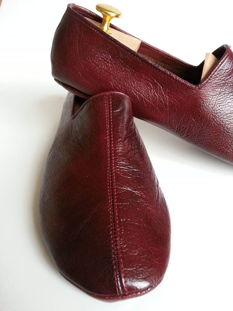 May include: A pair of burgundy leather slippers with a pointed toe and a stitched seam down the center. The slippers are made of a soft, supple leather and have a comfortable fit.