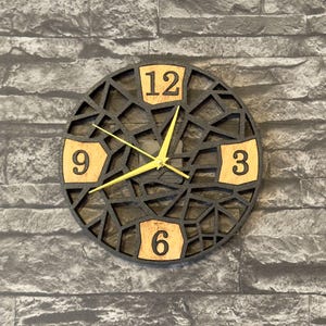 Könnte beinhalten: Eine runde, schwarze Wanduhr mit geometrischem Design. Das Zifferblatt zeigt die Zahlen 3, 6, 9 und 12 in Hellbraun. Die Zeiger der Uhr sind gelb.