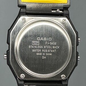 Pode incluir: Close-up de um rel&oacute;gio digital Casio F-94W. O rel&oacute;gio tem uma caixa octogonal preta com uma parte traseira em a&ccedil;o inoxid&aacute;vel e uma pulseira amarela. O mostrador do rel&oacute;gio exibe o texto "CASIO", "1071 F-94W", "STAINLESS STEEL BACK", "WATER RESISTANT", "MADE IN CHINA" e "DH".