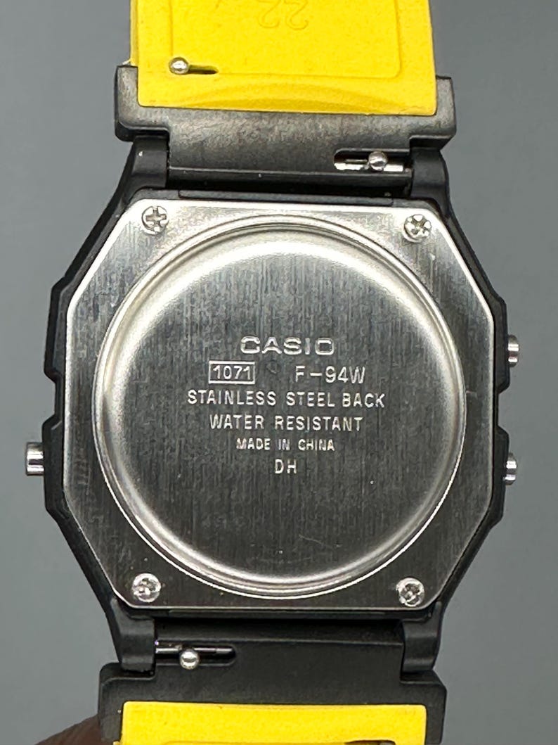 Pode incluir: Close-up de um rel&oacute;gio digital Casio F-94W. O rel&oacute;gio tem uma caixa octogonal preta com uma parte traseira em a&ccedil;o inoxid&aacute;vel e uma pulseira amarela. O mostrador do rel&oacute;gio exibe o texto "CASIO", "1071 F-94W", "STAINLESS STEEL BACK", "WATER RESISTANT", "MADE IN CHINA" e "DH".