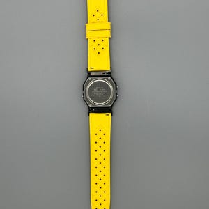 Pode incluir: Um rel&oacute;gio digital preto com uma pulseira amarela brilhante perfurada. O mostrador do rel&oacute;gio &eacute; quadrado e a pulseira &eacute; presa com uma fivela prateada. O rel&oacute;gio &eacute; exibido em um fundo cinza.